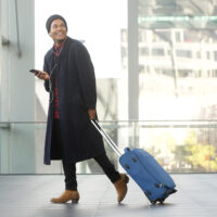 Full length smiling travel man walking at airport with cellphone and luggage
