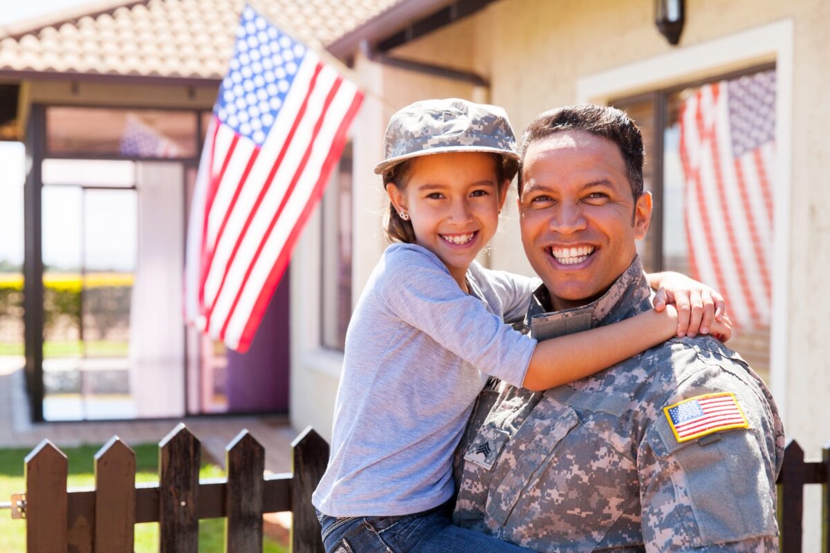 United States army soldier holding young daughter, standing near an American flag.